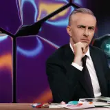 Mann mit nachdenklichem Blick hinter einem Schreibtisch, im Hintergrund eine abstrakte, bunte Wand und ein Fernglas mit seltsamen Symbolen, die wie gibberish language wirken.