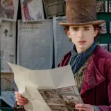 Timothée Chalamet als junger Willy Wonka, der in einem viktorianischen Outfit und mit einem Zylinder vor einem Zeitungskiosk steht und eine Broschüre liest. Die Besetzung von Wonka verspricht Magie.