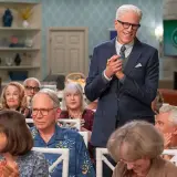 Ein Mann im Anzug steht vor einer Gruppe älterer Menschen, die applaudieren. Es sieht aus wie eine **Besetzung von undercover im Seniorenheim**.