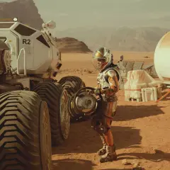 Ein Astronaut in einem orangefarbenen Raumanzug steht neben einem Mars-Rover. Szene aus dem Marsianer Film.