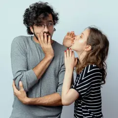 Sie flüstert ihm etwas ins Ohr, sein Gesichtsausdruck deutet auf ein **double entendre** hin.