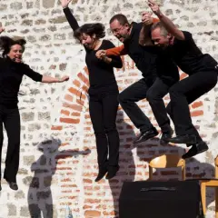 Vier Darsteller springen synchron in die Luft. Sie tragen schwarze Kleidung und Mikrofone. Ihre Mimik zeigt Freude und Energie, fast wie bei einem Playback-Auftritt.