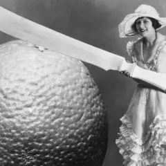 Eine Frau im Rüschenkleid hält ein riesiges Messer neben einer gigantischen Orange. Die Szene wirkt fast grotesk, eine humorvolle Darstellung von Größenverhältnissen.