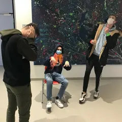 Drei Jungen mit Masken vor einem abstrakten Gemälde. Einer sitzt, die anderen stehen. Ein Moment wie ein Standbild im Theater.