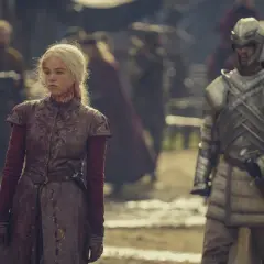 Rhaenyra Targaryen, blutverschmiert, blickt nachdenklich, während ein Ritter in voller Rüstung im Hintergrund der Folgen von House of the Dragon steht.