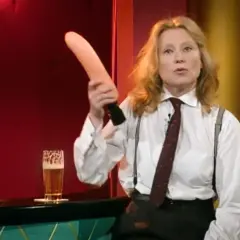 Eine Frau in einem weißen Hemd mit Krawatte und Hosenträgern hält einen rosafarbenen Gegenstand hoch. Was ist Sketch? Ein Glas Bier steht daneben.