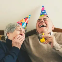 Ein älteres Paar mit Partyhüten lacht herzhaft. Sie zeigen, wie man jemanden zum Lachen bringt: mit Freude und Humor.
