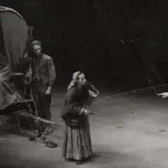 Szene aus einem **Brecht Theater**: Eine Frau mit Kopftuch und Korb steht vor einem Wagen. Zwei Männer in Rüstung zeigen in eine Richtung.