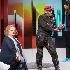 Ein Mann mit roter Kappe und Werkzeuggürtel reicht einer sitzenden Frau eine Sprühflasche. Ein Standbild-Beispiel einer Theateraufführung.
