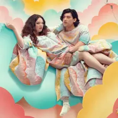 Ein Paar sitzt auf einer Wolke, eingehüllt in eine bunte Decke. Die besetzung von Too Much scheint in einer surrealen Traumwelt zu schweben.