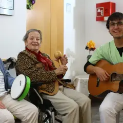 Zwei ältere Damen und ein junger Mann musizieren gemeinsam. Die Damen spielen Trommel und Maracas, der Mann spielt Gitarre. Ein fröhlicher Moment, wie im **Coco Film**.