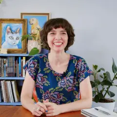 Eine Frau mit einem breiten Lächeln sitzt vor einem Bücherregal mit Katzen- und Hundebildern. Sie wirkt aufgeregt und lustig, bereit für ein Gespräch.