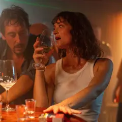 Ein Mann und eine Frau sitzen an einer Bar, sie trinkt aus einem Weinglas. Die Szene erinnert an den Film "der rausch film", mit gedämpftem Licht und einer intimen Atmosphäre.
