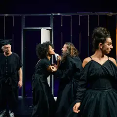 Schauspieler in Schwarz auf einer Bühne, ein Mann mit Hut, ein Paar im Streit, eine Frau im Vordergrund. Ein **open mic** Moment voller Dramatik.