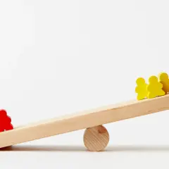 Eine rote Figur steht allein auf einer Seite einer Wippe, während drei gelbe Figuren auf der anderen Seite sind. Die Wippe ist geneigt, was hochstatus tiefstatus symbolisiert.