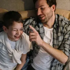 Ein Vater erzählt seinem Sohn einen Knock-Knock-Witz. Der Junge lacht herzhaft, während der Vater ihm mit einem Finger deutet.