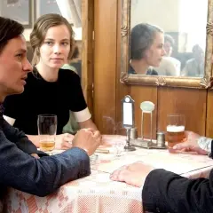Fabian oder der Gang vor die Hunde: Ein Mann mit Bart spricht mit einer Frau, während ein anderer Mann im Vordergrund sitzt.