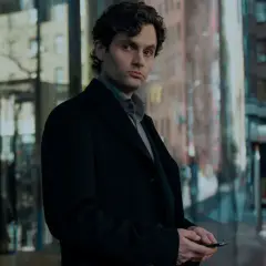 Joe Goldberg aus der serie "You" steht nachdenklich in Schwarz gekleidet vor einem modernen Gebäude.