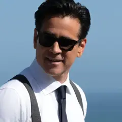 John Sugar, mit Sonnenbrille und Hosenträgern, lächelt vor dem blauen Himmel und Meer.