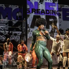 Künstler mit Afro-Perücke singt in glitzerndem Outfit. Im Hintergrund tanzen Menschen, einige in orangefarbenen Overalls. Was ist Monolog? Eine Performance, die fesselt.