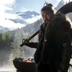Mann mit Fellmantel und Axt blickt ernst in die Kamera, im Hintergrund Berge und Wald. Ein Feuer brennt in einem Kessel. Sieh diese Serie!