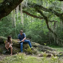 Ein Mann und eine Frau sitzen unter einem Baum im Wald. Szene aus dem Film "Der Gesang der Flusskrebse".