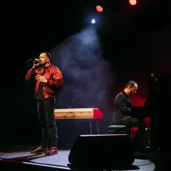 Ein Sänger mit Mikrofon und ein Pianist auf der Bühne. Die Beleuchtung schafft eine dramatische Atmosphäre, die gut zu spontanen **Stegreifrede Themen** passt.