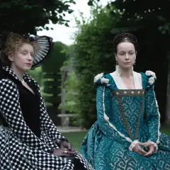 Zwei Frauen in historischen Gewändern, eine in Schwarz-Weiß, die andere in Türkis, sitzen im Garten. Die eine könnte die Schlangen-Königin sein, die andere ihre Rivalin.