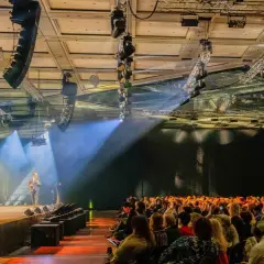 Bühne mit Redner, Publikum und Lichtshow. Die Atmosphäre erinnert an eine spontane Improvisation, Musik und ein inspirierendes Event.