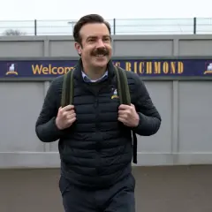 Ted Lasso, mit seinem charakteristischen Schnurrbart, trägt einen Rucksack vor dem Schild "Welcome to Richmond".