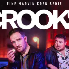 Zwei Männer, Teil der "Crooks"-Besetzung, stehen im nächtlichen Stadtlicht. Einer hält eine Münze, der andere blickt nachdenklich.