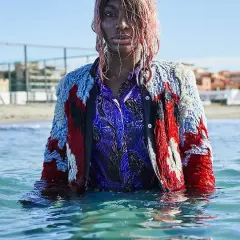 Ein Model mit pinken Haaren steht im Meer, trägt eine bunte Jacke und ein blaues Hemd. Die Wellen umspielen sie, als ob sie sagen: "I may destroy you.