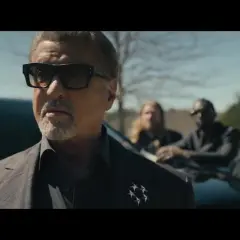 Sylvester Stallone als Tulsa King, mit Sonnenbrille und Anzug, steht vor einem Auto, begleitet von drei Männern.