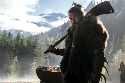 Mann mit Fellmantel und Axt blickt ernst in die Kamera, im Hintergrund Berge und Wald. Ein Feuer brennt in einem Kessel. Sieh diese Serie!
