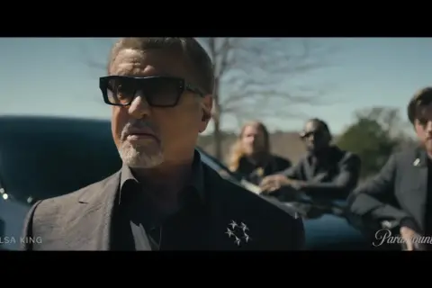 Sylvester Stallone als Tulsa King, mit Sonnenbrille und Anzug, steht vor einem Auto, begleitet von drei Männern.