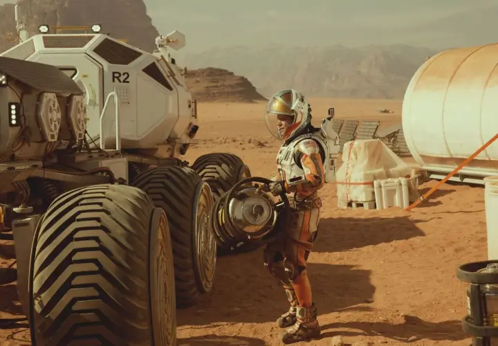 Ein Astronaut in einem orangefarbenen Raumanzug steht neben einem Mars-Rover. Szene aus dem Marsianer Film.