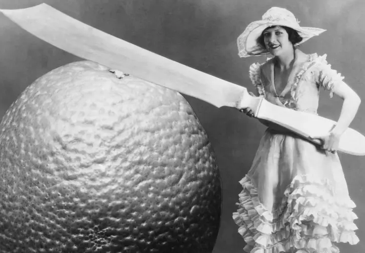 Eine Frau im Rüschenkleid hält ein riesiges Messer neben einer gigantischen Orange. Die Szene wirkt fast grotesk, eine humorvolle Darstellung von Größenverhältnissen.