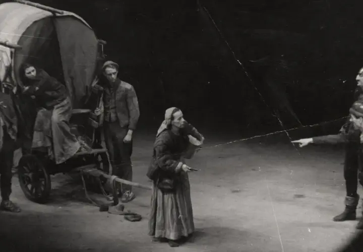Szene aus einem **Brecht Theater**: Eine Frau mit Kopftuch und Korb steht vor einem Wagen. Zwei Männer in Rüstung zeigen in eine Richtung.