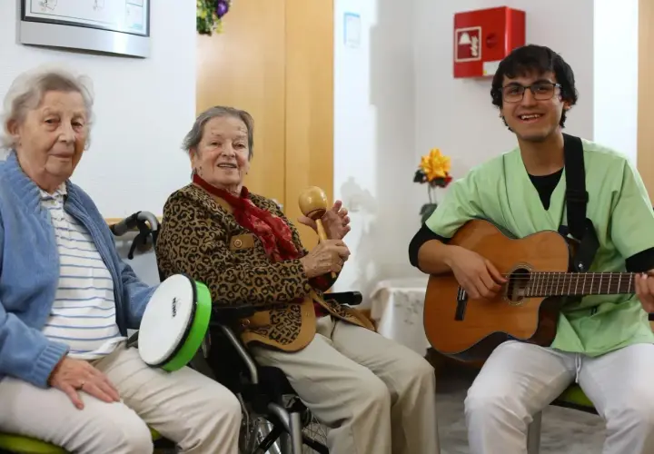 Zwei ältere Damen und ein junger Mann musizieren gemeinsam. Die Damen spielen Trommel und Maracas, der Mann spielt Gitarre. Ein fröhlicher Moment, wie im **Coco Film**.