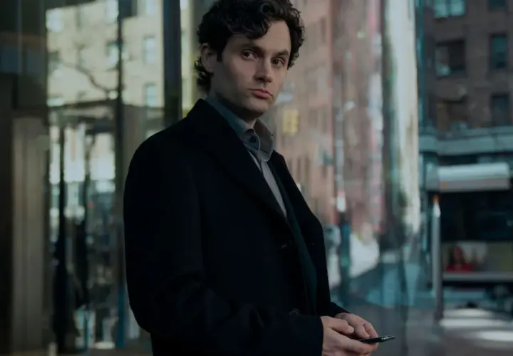 Joe Goldberg aus der serie "You" steht nachdenklich in Schwarz gekleidet vor einem modernen Gebäude.