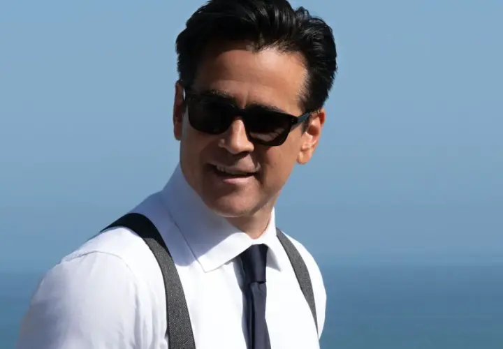 John Sugar, mit Sonnenbrille und Hosenträgern, lächelt vor dem blauen Himmel und Meer.