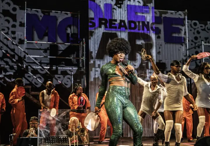 Künstler mit Afro-Perücke singt in glitzerndem Outfit. Im Hintergrund tanzen Menschen, einige in orangefarbenen Overalls. Was ist Monolog? Eine Performance, die fesselt.