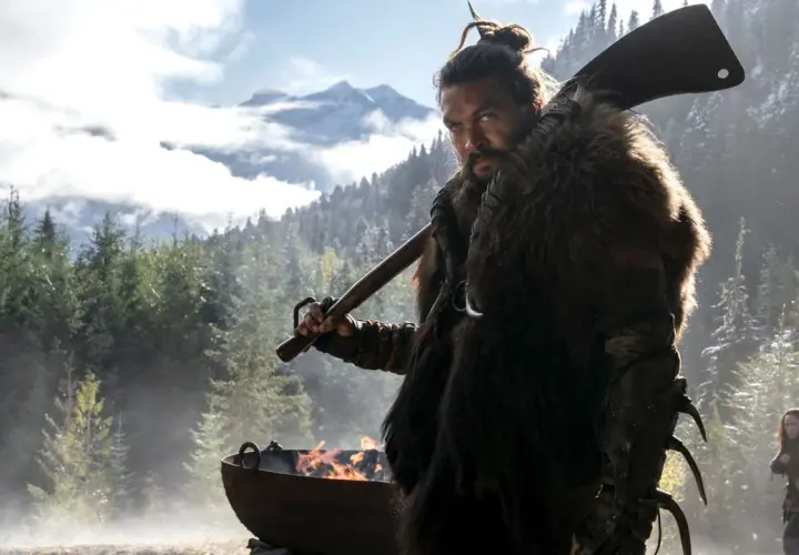 Mann mit Fellmantel und Axt blickt ernst in die Kamera, im Hintergrund Berge und Wald. Ein Feuer brennt in einem Kessel. Sieh diese Serie!