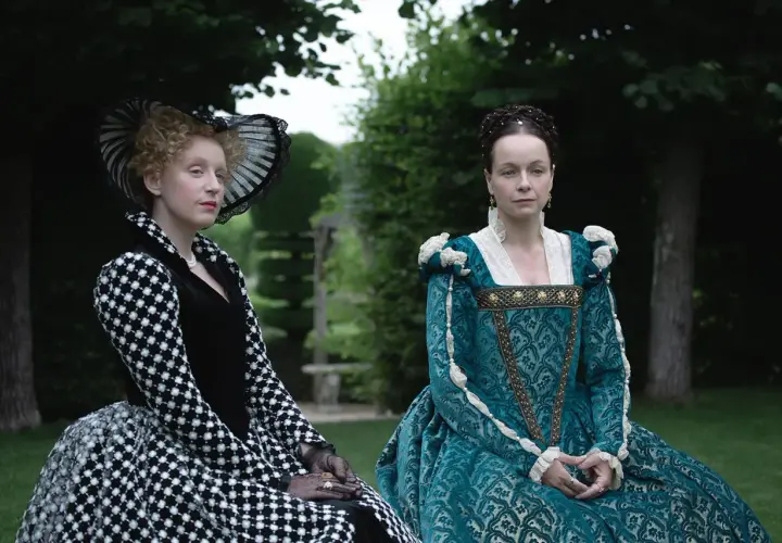 Zwei Frauen in historischen Gewändern, eine in Schwarz-Weiß, die andere in Türkis, sitzen im Garten. Die eine könnte die Schlangen-Königin sein, die andere ihre Rivalin.
