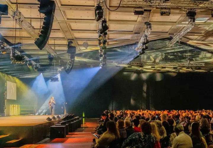 Bühne mit Redner, Publikum und Lichtshow. Die Atmosphäre erinnert an eine spontane Improvisation, Musik und ein inspirierendes Event.