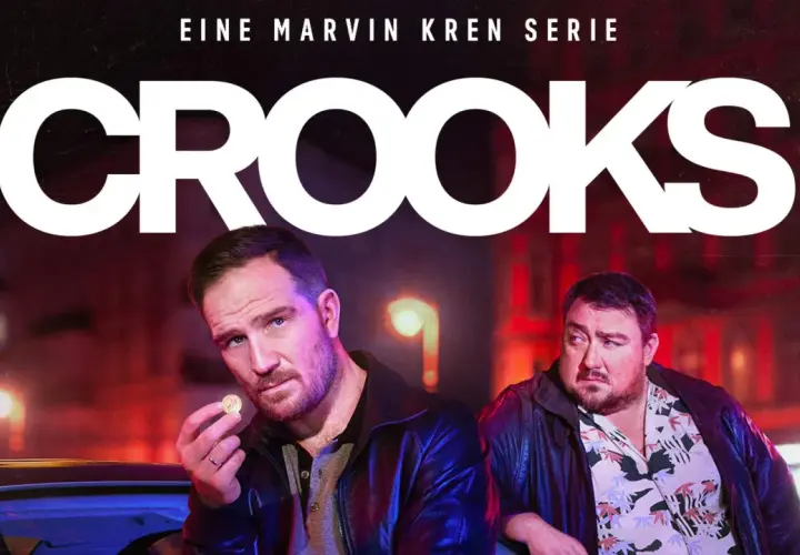 Zwei Männer, Teil der "Crooks"-Besetzung, stehen im nächtlichen Stadtlicht. Einer hält eine Münze, der andere blickt nachdenklich.