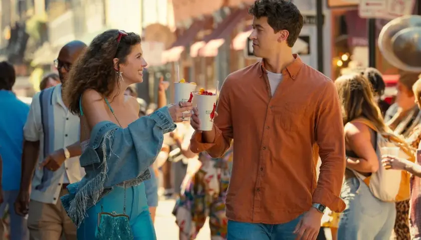 Ein junges Paar stößt mit Getränken an, während es durch eine belebte Straße schlendert. Die Besetzung von "People We Meet on Vacation" scheint hier ihren Urlaub zu genießen.