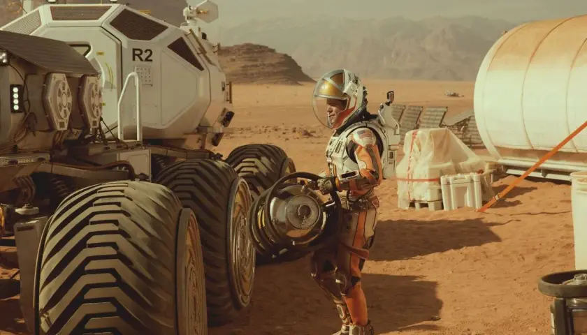 Ein Astronaut in einem orangefarbenen Raumanzug steht neben einem Mars-Rover. Szene aus dem Marsianer Film.