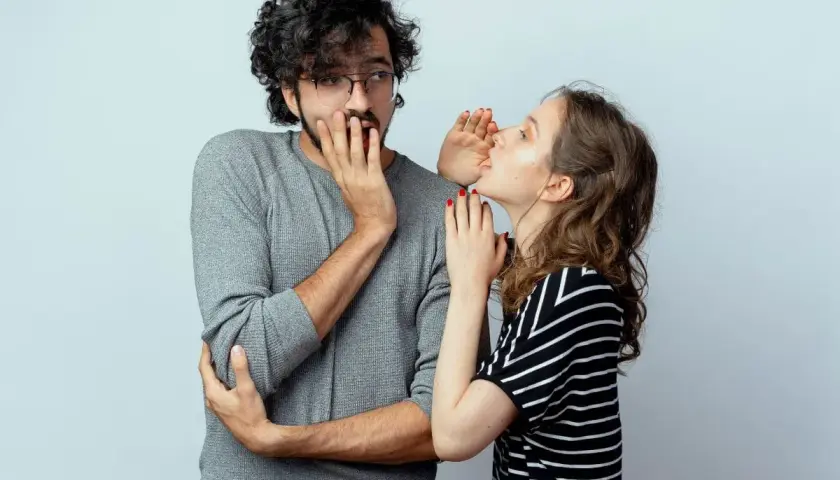 Sie flüstert ihm etwas ins Ohr, sein Gesichtsausdruck deutet auf ein **double entendre** hin.