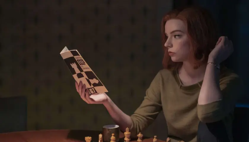 Beth Harmon studiert eine Schachpartie, inspiriert vom Damengambit. Schachfiguren und eine Tasse stehen auf dem Tisch.